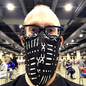 A selfie taken by the author, smiling broadly behind a large mask covering a large beard, sitting in a chair in the concrete floor of a cavernous convention center. Other people can be seen in the background also sitting on socially distanced chairs.