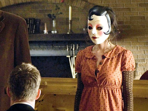 A still from The Strangers, with a woman in a “pin-up doll” mask facing a seated man