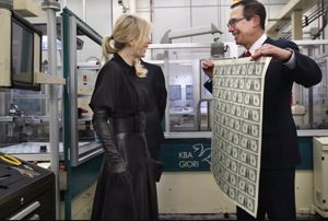 Louise Linton standing with Steve Mnuchin, who is holding an uncut sheet of freshly-printed $1 bills. Linton’s all-black outfit evokes Darth Vader