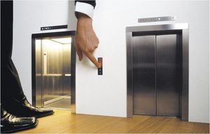 A man in a business suit presses the button to call an elevator that is much too small for him. It’s unclear if he is huge or the elevator is tiny. It’s also unclear why he’s pressing the button when the elevator is already waiting for him with the door open. The parts of him in frame seem to be impossibly contorted.