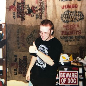 A doofus in a Gwar t-shirt giving a thumbs-up in front of a pair of curtains fashioned from burlap sacks and a mini fridge with a “BEWARE OF DOG” sign on the door.