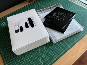 A stack of white paperboard mailers with address labels, sitting on a green cutting mat next to a pile of three small, square, paperback books with a large white 6 on their black covers